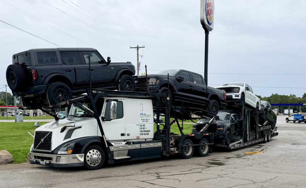 Truck carrier with a white Volvo cab and multi-level car hauler loaded with several black trucks.
