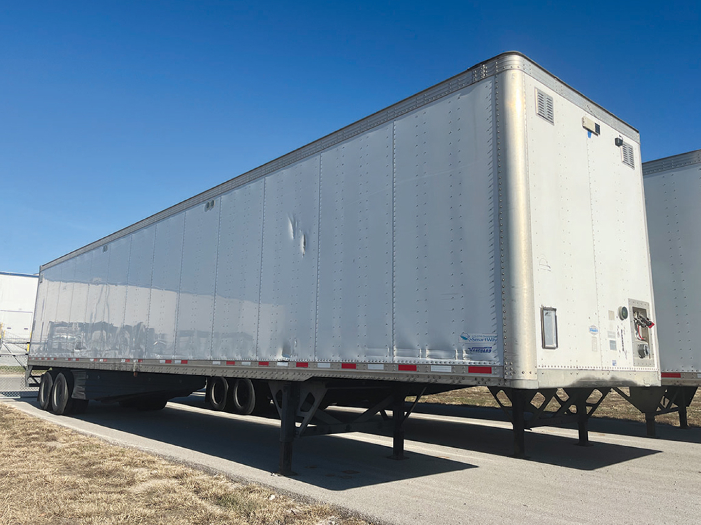 Trailer image showing a long silver enclosed cargo trailer with dual rear axles, elevated hitch, and side riveted panels.