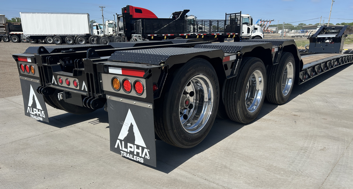Heavy-duty lowboy trailer with three rear axles showing dual mud flaps with Alpha Trailers logo, taillights, and reflective strips; aluminum wheels and black metal frame; trailing end of trailer.