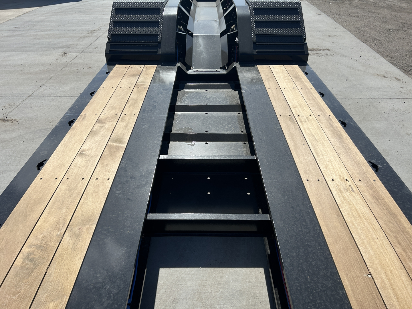 Lowboy trailer deck showing wooden planks on both sides with black steel frame and gooseneck area at the far end; no visible labels or text.