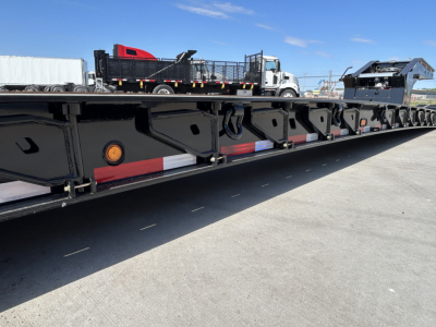 Lowboy trailer showing side rails and deck with cutouts and reflectors; machinery and vehicles on a flatbed in background.
