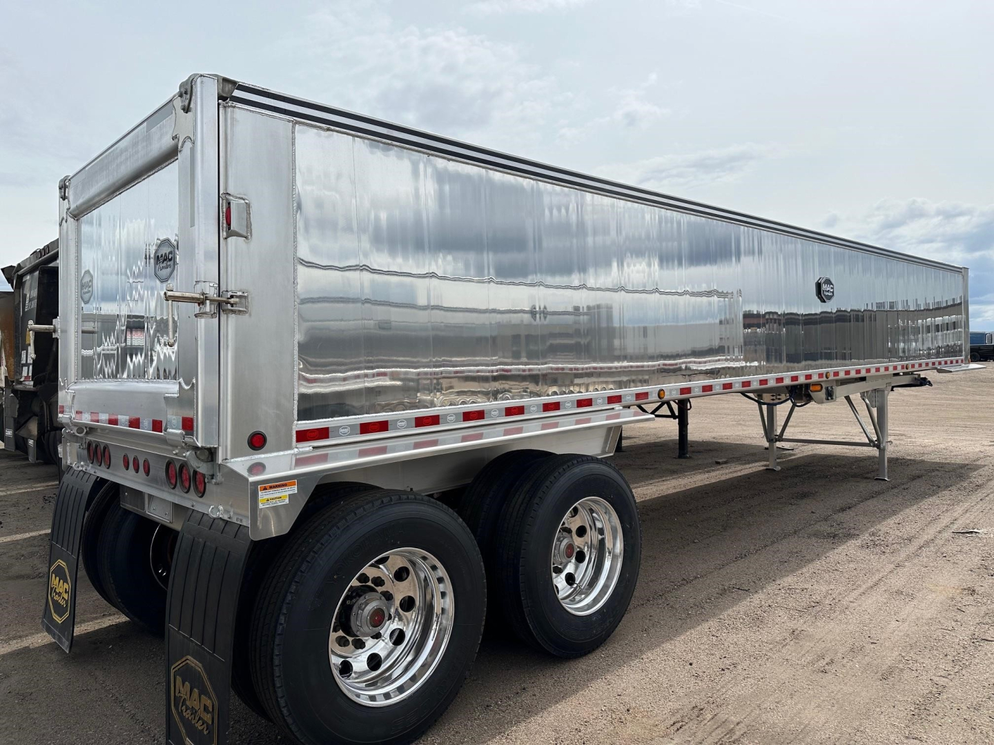 End dump trailer with polished aluminum finish showing dual rear axles and chrome wheels; side panels and rear gate visible.