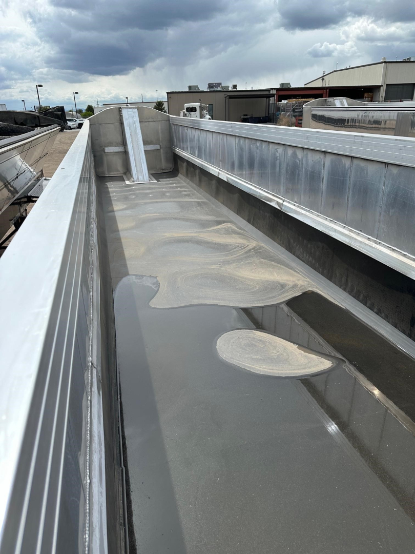 Truck trailer bed showing aluminum end dump with reflective side rails and a shallowly curved bed. Visible on the bed are circular light tan deposits and metal surfaces.