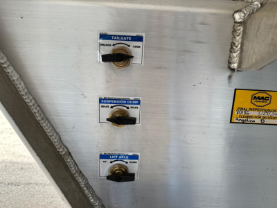 Mac trailer end-dump control panel with three levers labeled 'TAILGATE', 'SUSPENSION DUMP', and 'LIFT ARLE' on a metal surface; small stickers and welded seams visible.