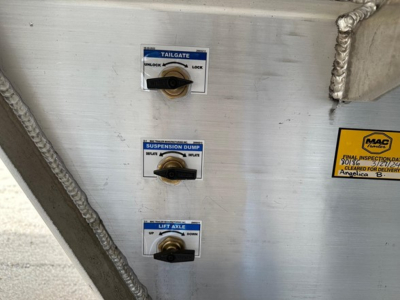 Trailer controls panel with three blue-labeled levers: Top labeled 'TAILGATE' with positions LOCK/UNLOCK; middle labeled 'SUSPENSION DUMP' with DOWN/UP; bottom labeled 'LIFT AXLE' with DOWN/UP. Small MAC trailer sticker on right.