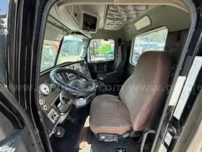 Cab interior of a 2015 Freightliner Coronado truck showing steering wheel, driver seat, dashboard with gauges, overhead compartments, and front windows.