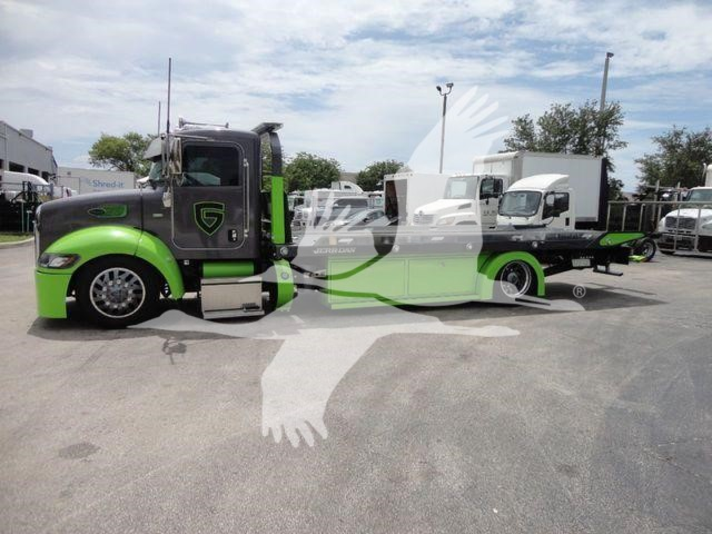 2020 Peterbilt 337 rollback truck in a lot, front cab is green and black, low-profile sleeper, long wheelbase with a green undercarriage, logo on door, various equipment and trailers in background.
