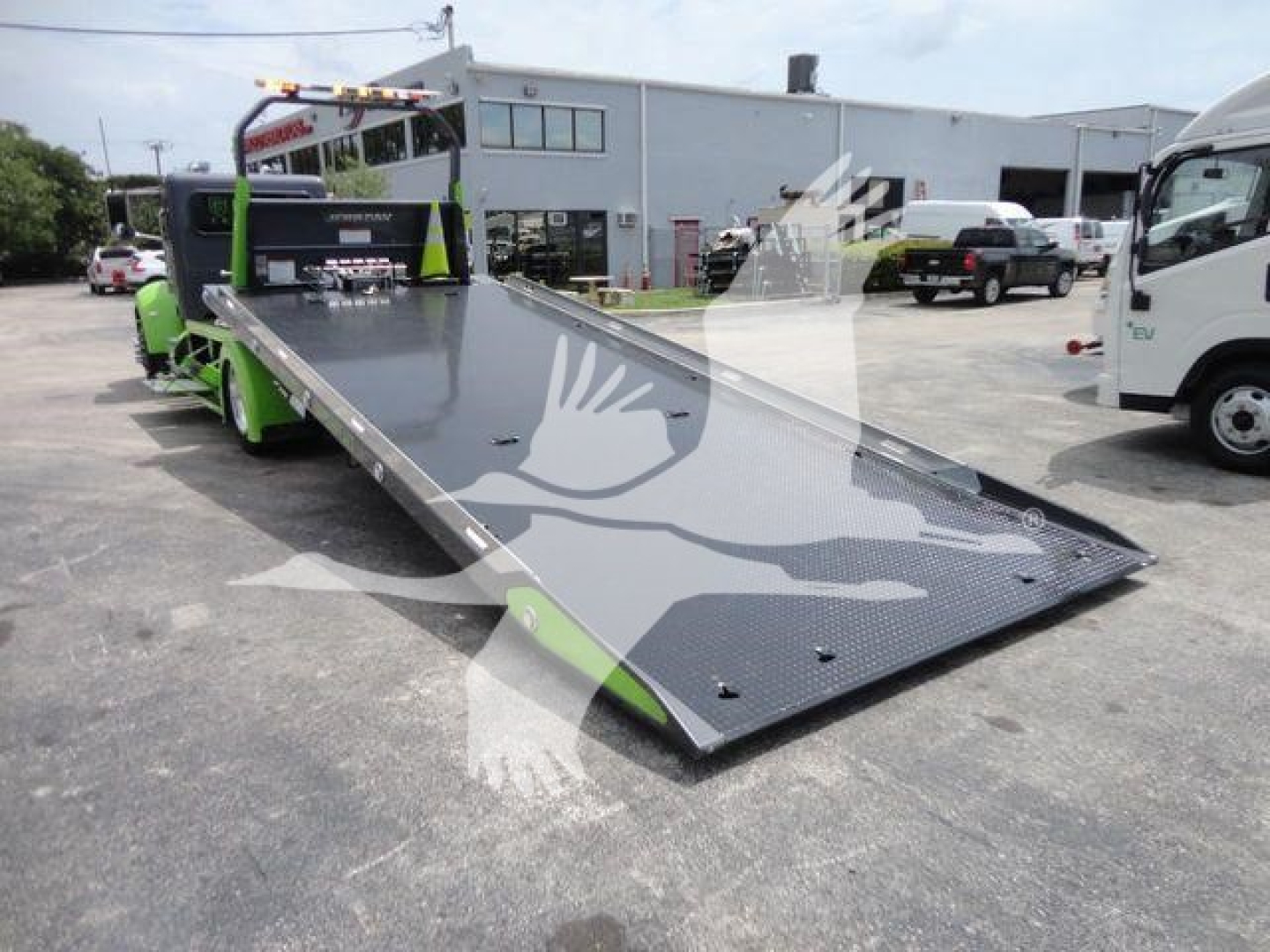 Peterbilt 337 rollback truck with a lowered flatbed, green accents, and ramp in foreground. Side view shows cab and yard with other trucks.