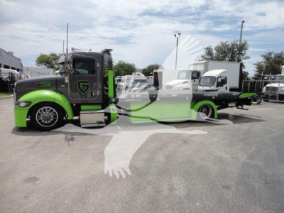 2020 Peterbilt 337 rollback truck in a lot, front cab is green and black, low-profile sleeper, long wheelbase with a green undercarriage, logo on door, various equipment and trailers in background.