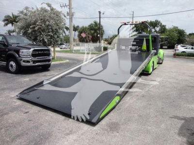 Green Peterbilt rollback tow truck with a long flatbed in a dealership lot, front of bed visible, other vehicles in background.