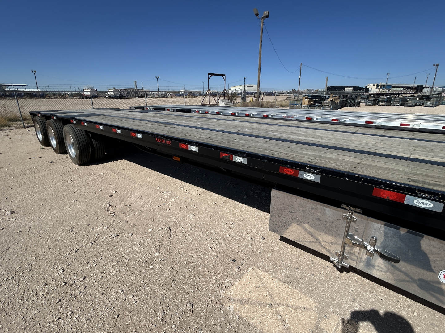 Flatbed trailer visible with dual rear axles in a gravel lot; long deck, chain storage racks along side; Clear weather.