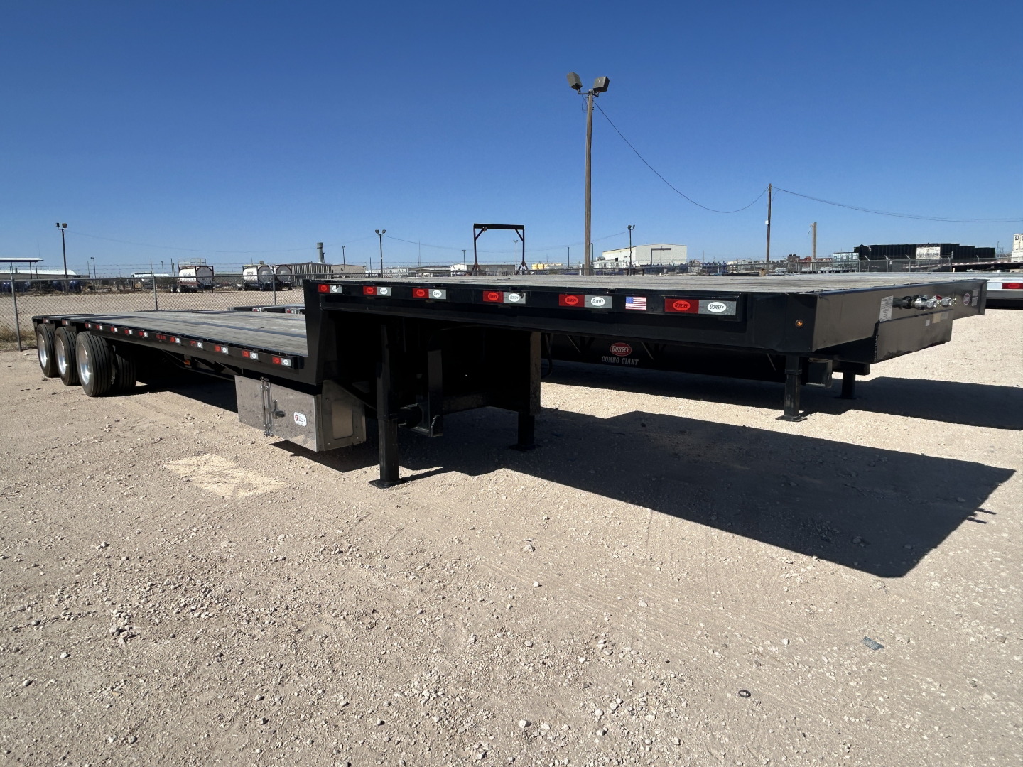 Trailer showing flatbed with dual rear axles and drop deck, black metal frame, with stakes and deck supports visible.