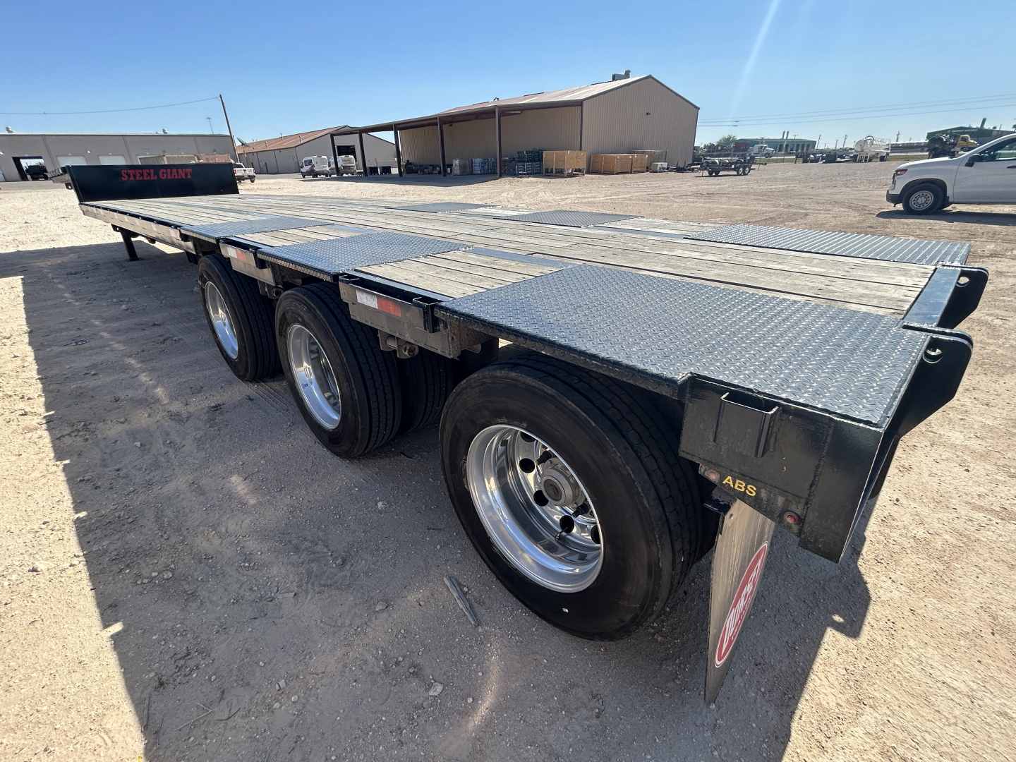 Flatbed trailer with triple-axle setup and diamond plate deck, side rails and rear ramps visible; metal frame and wheels.