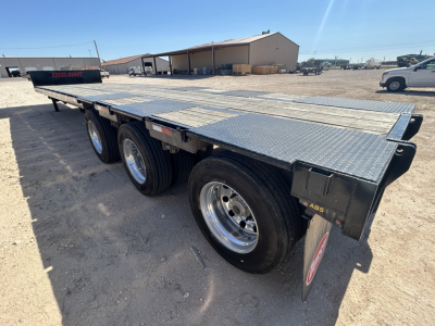 Flatbed trailer with triple-axle setup and diamond plate deck, side rails and rear ramps visible; metal frame and wheels.
