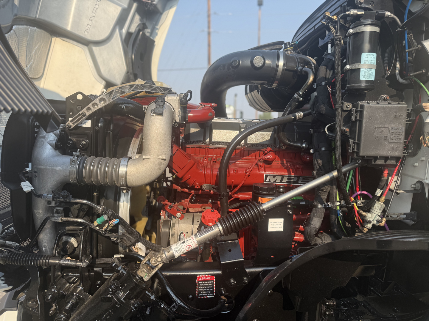 Truck engine bay showing Mack ANTHEM with red engine block, hoses, and various components; visible labels and wiring harnesses.