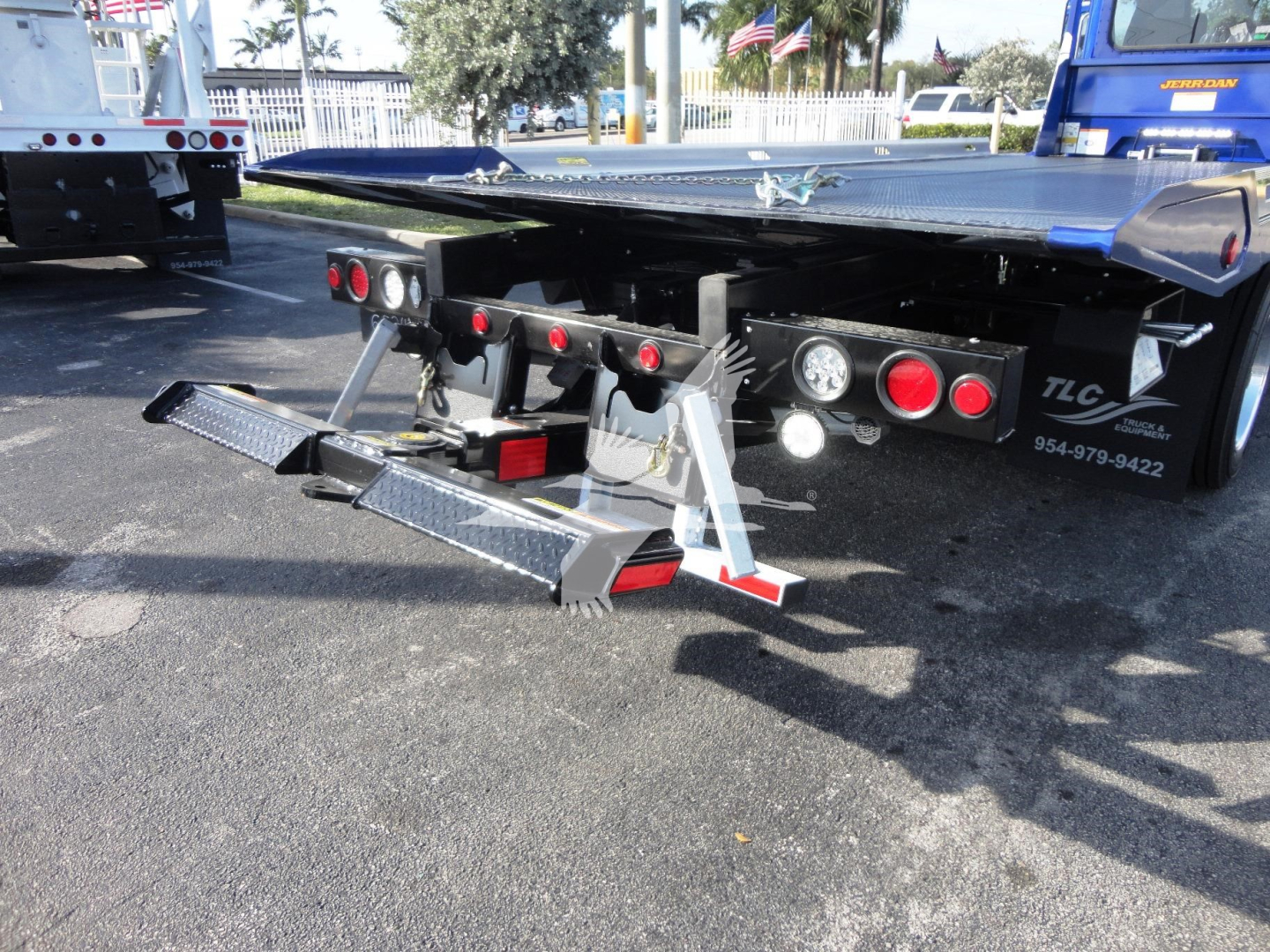 Peterbilt 337 rollback truck rear showing hydraulic ramps and taillights, stock number J32060 FM text on the chassis, TLC phone number 954-979-8422, and a blue truck in background.