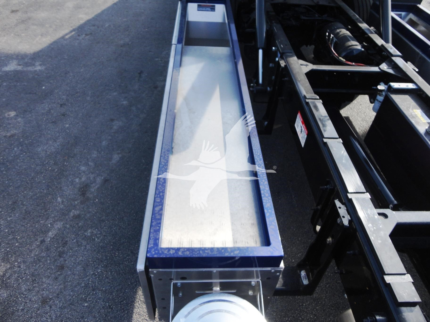 Truck component bay with metallic frame and a long rectangular trough; visible are hydraulic lines and a circular component at the end; no legible text readable.