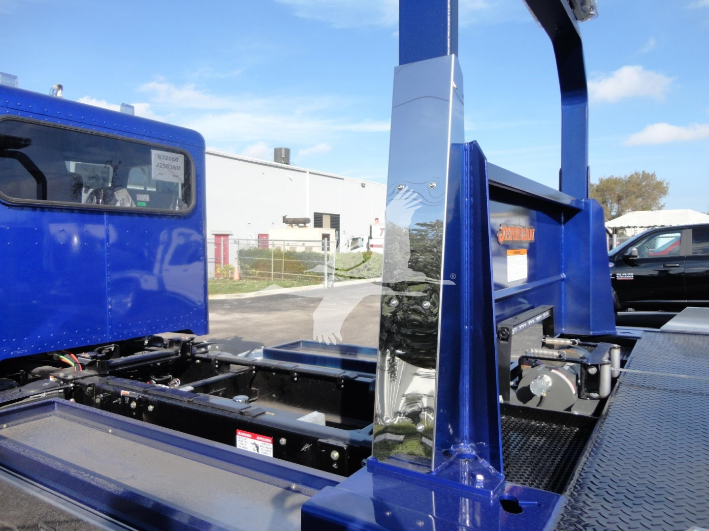 Truck chassis and controls showing blue Peterbilt rollback setup with metal platform, hydraulic arms, and side panels. Visible text includes a small sticker on the window and labels on the equipment.