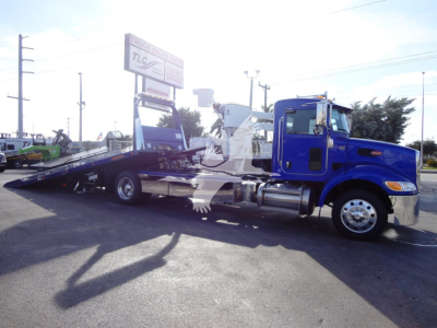 Blue 2022 Peterbilt 337 rollback wrecker chassis with black flatbed in a dealership lot showing long raised bed and chrome wheels.
