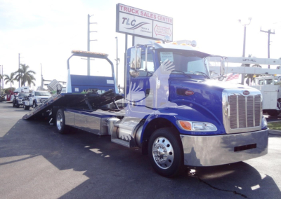 Peterbilt 337 rollback tow truck in blue with flatbed, chrome grille and bumper, parked at dealership lot.