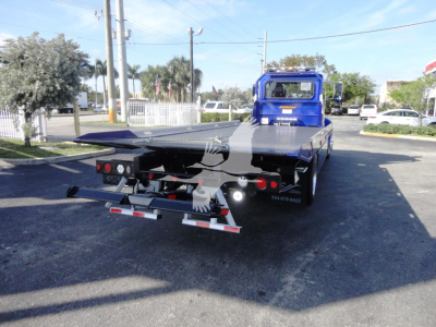 Back view of a blue Peterbilt 337 rollback tow truck with a flatbed, white and red safety markings on the rear, and light fixtures.