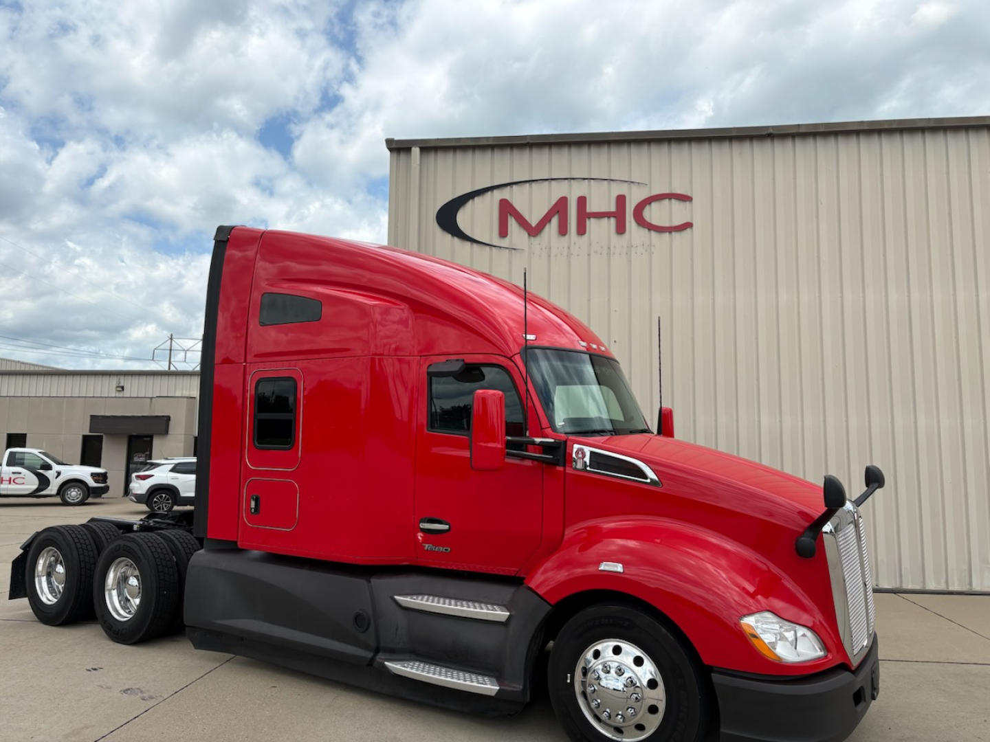 Kenworth T680 tractor in red with sleeper, side view showing front wheel and cab detail, dealership building in background with MHC logo.