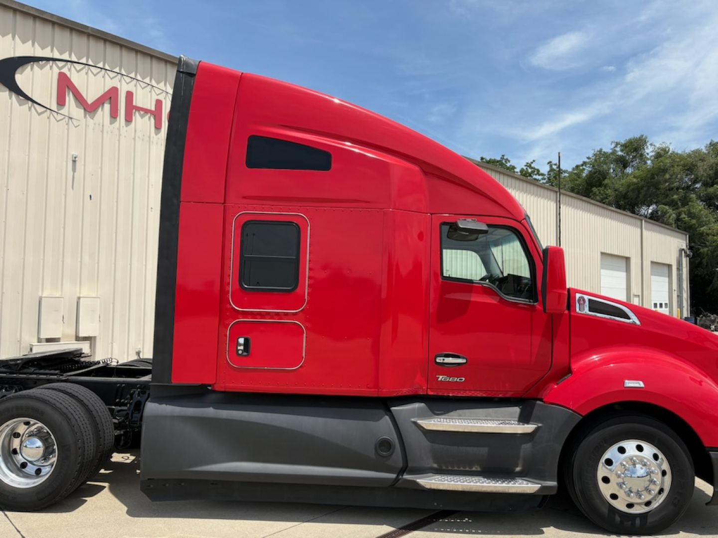 Kenworth T680 sleeper cab in red with aerodynamic hood, side door, and black lower body trim; truck on a lot with MH sign in background
