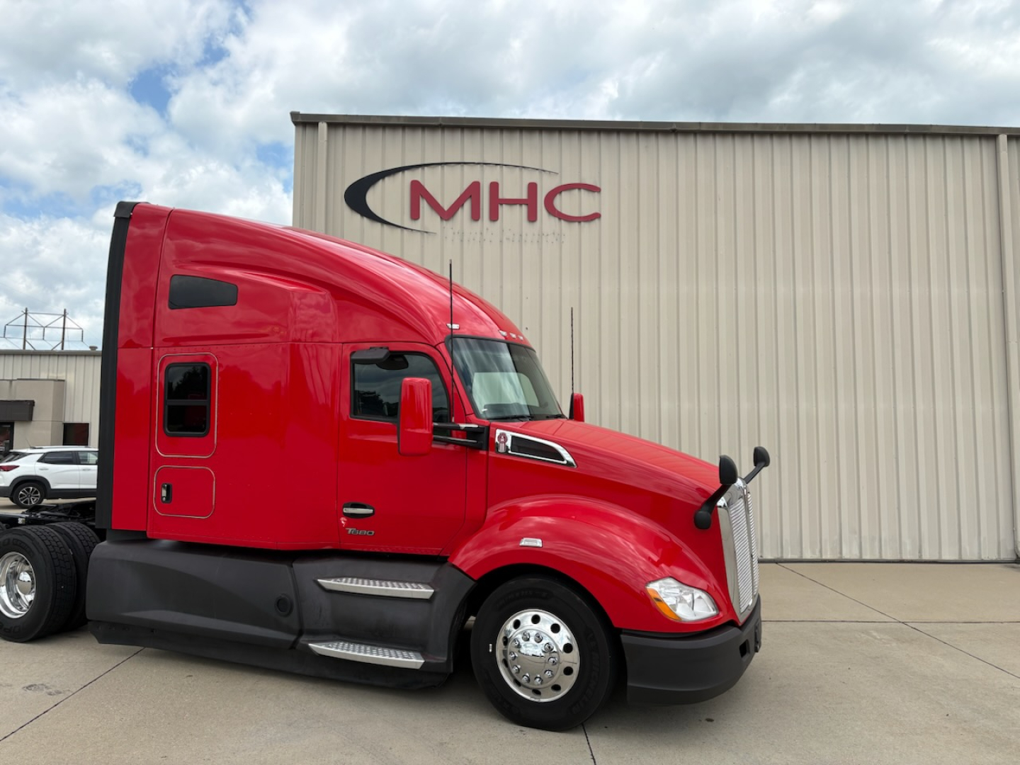 Kenworth T680 truck in red paint parked in front of MHC building; side profile with sleeper cab and chrome fuel tanks visible.