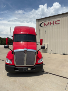 Kenworth T680 truck in red color facing forward showing bold grille and headlights; building with MHC logo in background; clear sky.