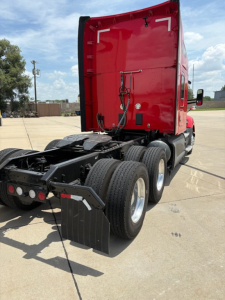Kenworth truck rear view showing red cab, dual rear axles, wheel mud flaps, and chassis with trailer air lines.