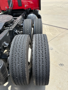 Kenworth truck rear wheel area showing dual rear axles with tires and suspension components; red cab in background.