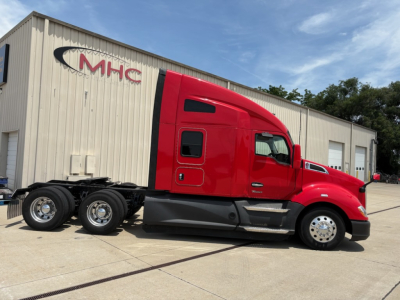 Kenworth T680 sleeper cab truck in red, side view with chassis and wheels, showroom/yard setting, MHC logo on building in background.