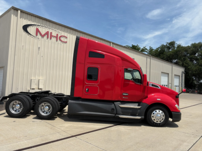 Side view of a red 2022 Kenworth T680 truck parked outside a maintenance facility with a long cab and sleeper, showing the wheelset and chrome wheels.