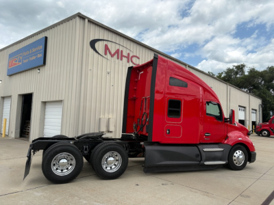 Truck cab exterior side view showing red Kenworth T680 with black chassis and triple rear axles, building with MHC sign in background.