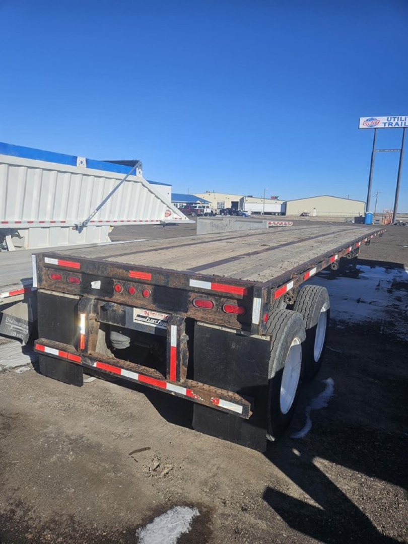 Flatbed trailer rear view showing dual rear axles and metal decking. Tail lights, reflective tape, and mud flaps visible. No explicit model numbers readable.