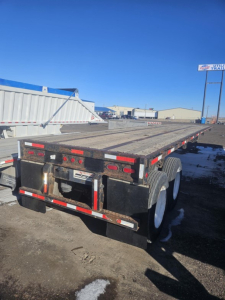 Flatbed trailer rear view showing dual rear axles and metal decking. Tail lights, reflective tape, and mud flaps visible. No explicit model numbers readable.