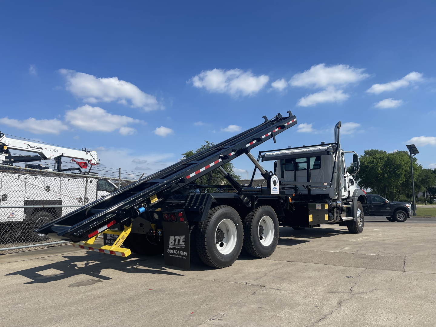 New 2027 Mack GR64B roll-off truck with dual rear axles and a tilted bed in a lot.