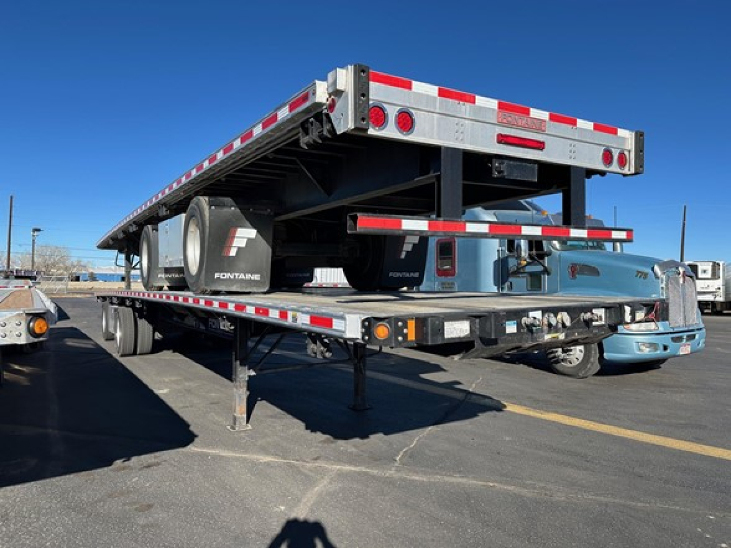 Flatbed trailer in-view showing Fontaine Infinity logo, dock leveler, and rear lighting; side rails and undercarriage with spare wheel, on-loading in a dealership lot.