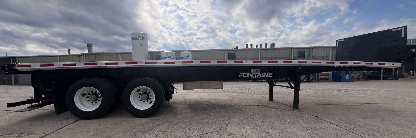 Flatbed trailer showing Fontaine logo with dual rear axles and fork lift kit visible, long aluminum deck, front landing gear, blue sky with clouds.