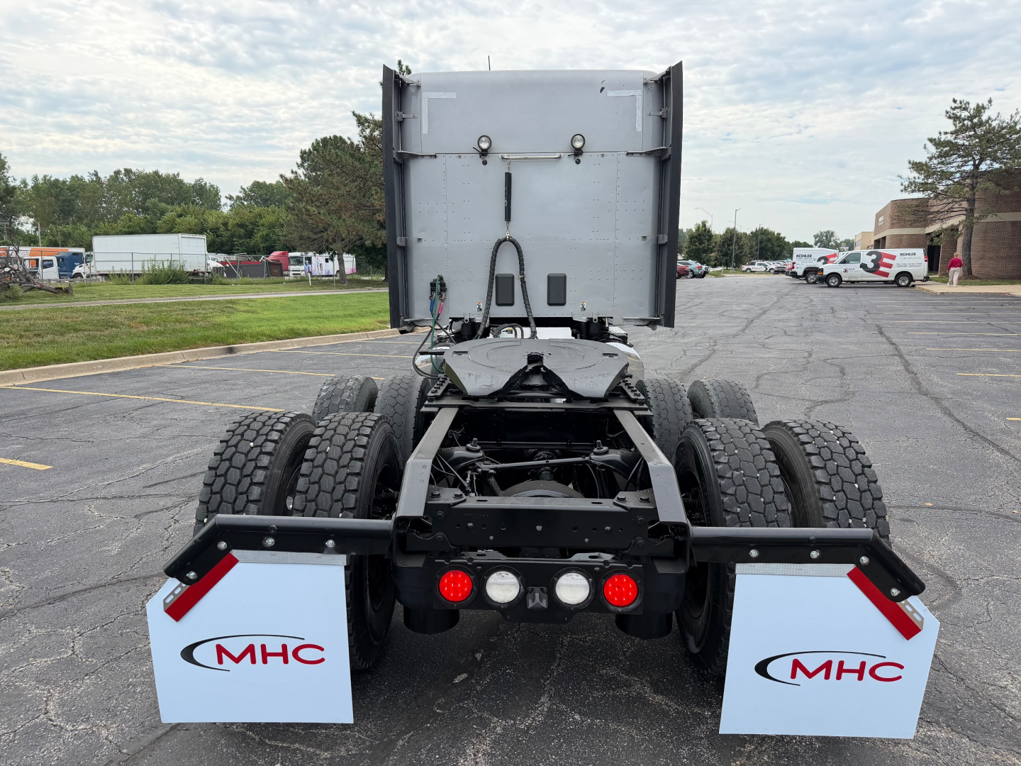 Truck rear view showing fifth-wheel attachment and mud flaps with MHC branding on a Kenworth T680 chassis. Dual rear axles with heavy-duty tires.