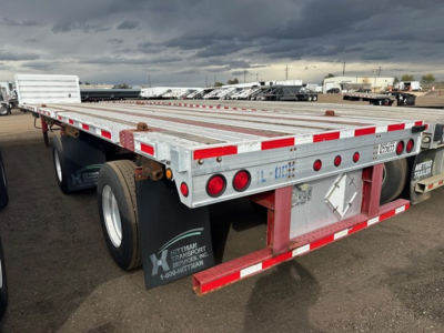 Flatbed trailer with metal decking and red-white reflective tape and rear lights, viewed from the left-rear side.