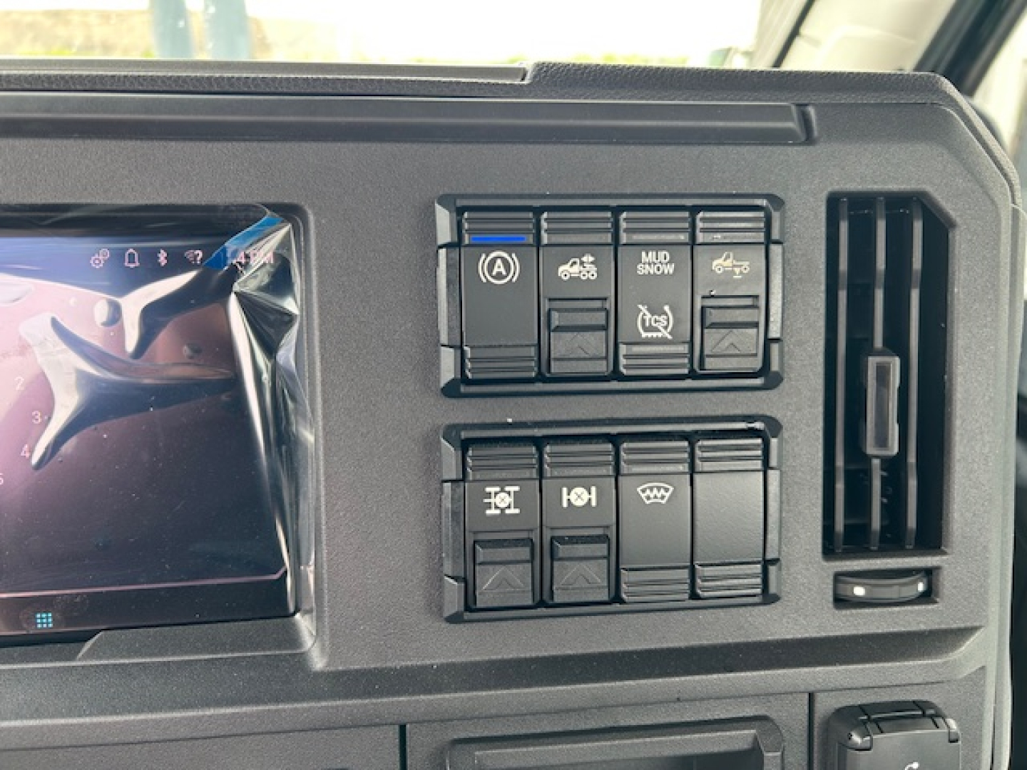Dashboard panel of a 2027 Mack AN64T showing multiple control switches and air vents with a screen on the left.