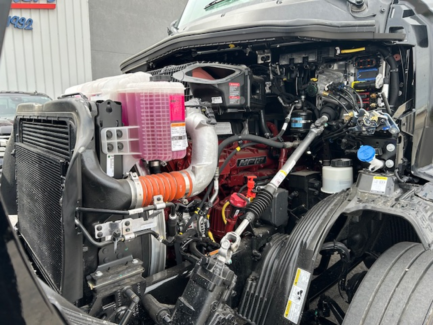 Front left engine bay of a new 2027 Mack AN64T daycab showing radiator, pink coolant reservoir, hoses, and various engine components visible