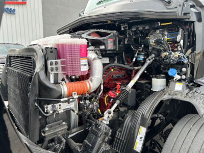 Front left engine bay of a new 2027 Mack AN64T daycab showing radiator, pink coolant reservoir, hoses, and various engine components visible