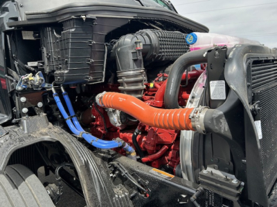 Truck engine bay view with orange exhaust/intercooler hose, blue hoses, red engine block, radiator, and surrounding components in a Mack AN64T cab/chassis.