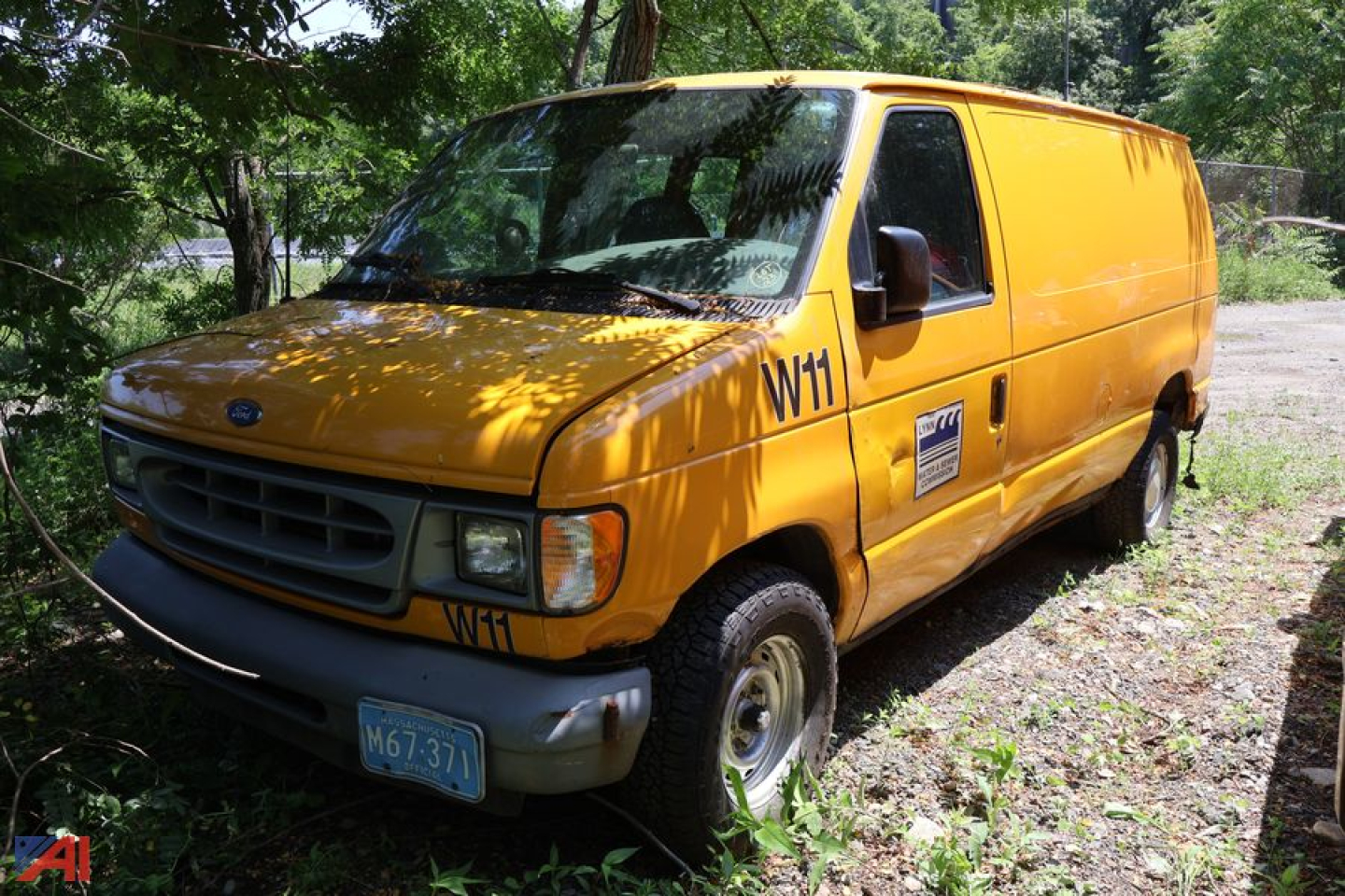 2002 Ford E150 cargo van in orange/yellow color, side door, front view with black grille and blue license plate. Marked WT on front fender and a small service sticker on the side.