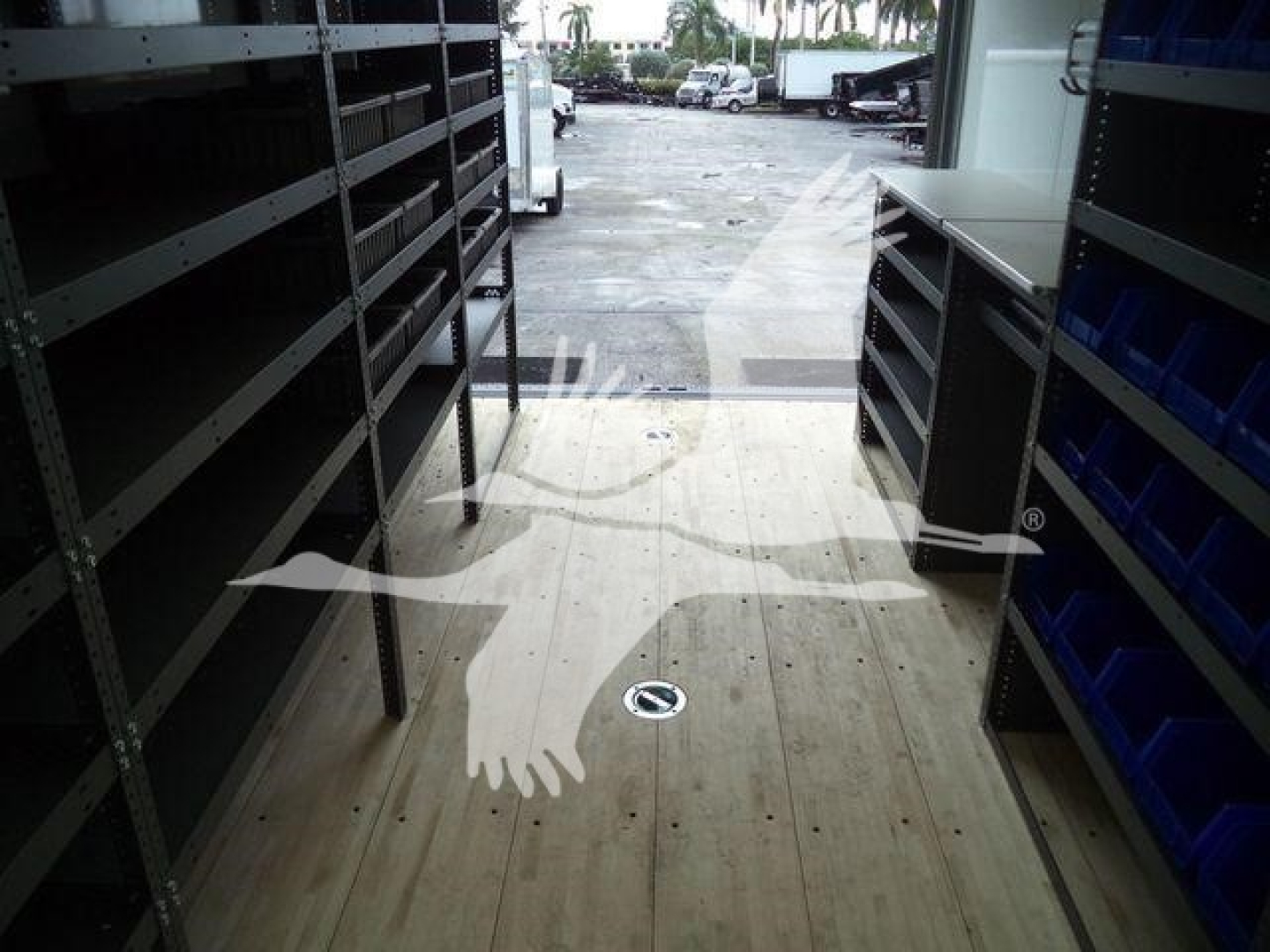 Truck showroom or trailer interior with shelves and blue bins, wooden floor, open doorway to exterior.