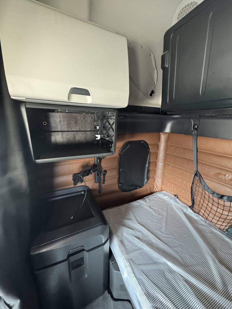 Cab interior of a Freightliner Cascadia 126 sleeper showing side storage, a small shelf, brown leatherette paneling, and a mattress with a light cover.