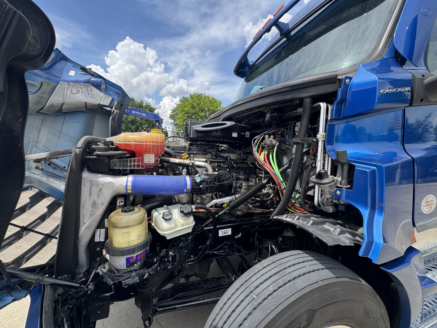 Truck engine bay of a blue 2022 Freightliner Cascadia 126 showing engine components, hoses, wiring, and reservoir caps.