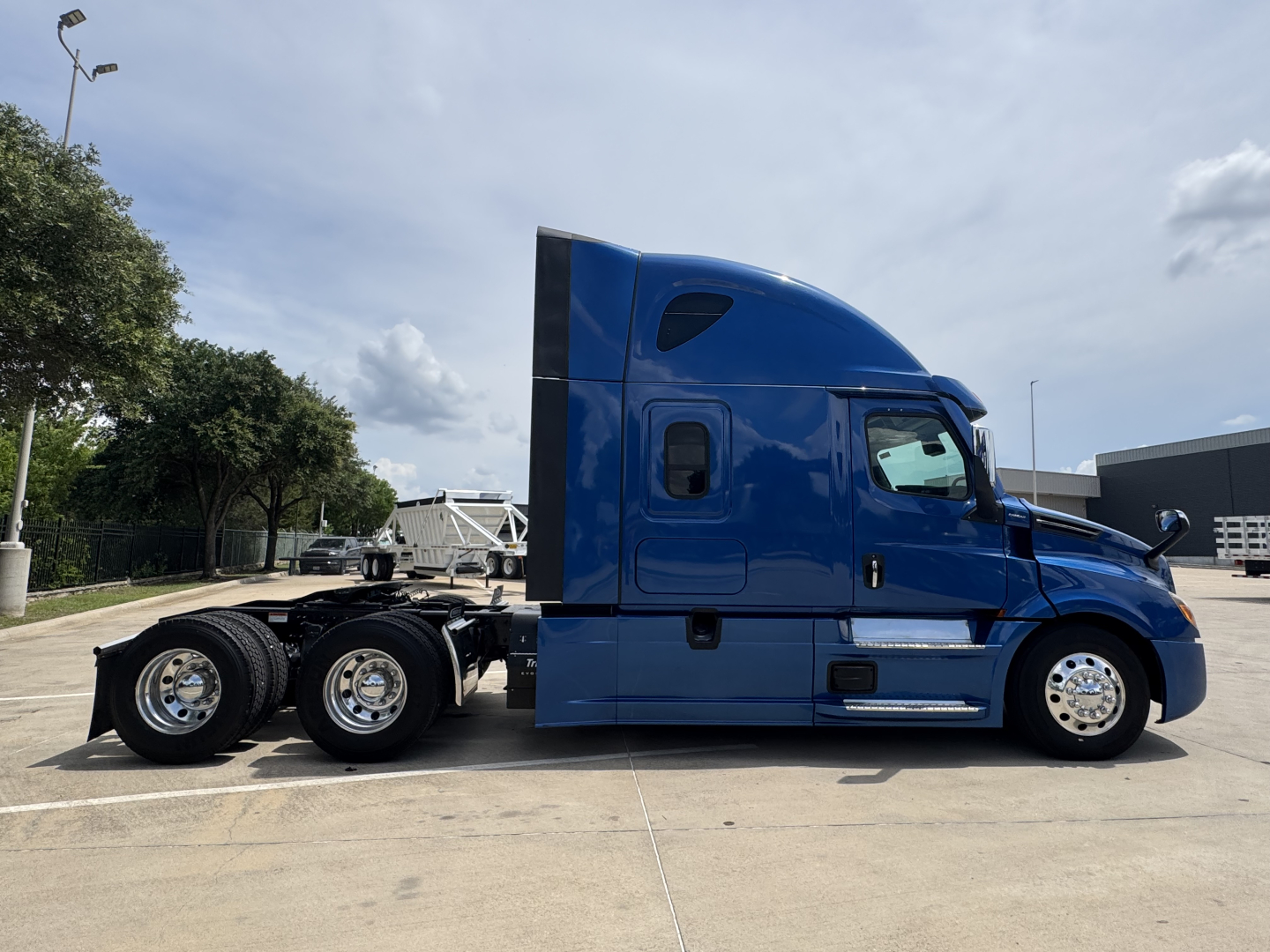 Side view of a blue 2022 Freightliner Cascadia 126 sleeper tractor in a parking lot, showing the cab, two rear axles, chrome wheels, and side mirrors.